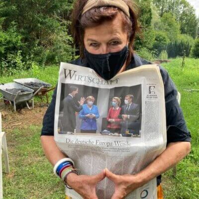Die Autorin Dodo Lazarowicz steht mit Maske und Wirtschaftsteil einer Zeitung in einem Garten.