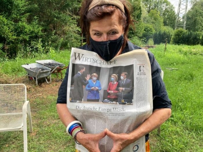 Die Autorin Dodo Lazarowicz steht mit Maske und Wirtschaftsteil einer Zeitung in einem Garten.