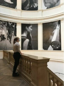 Eine Frau mit Mundschutz steht auf einer Empore in einem Museum, hinter ihr große Fotografien an der Wand.
