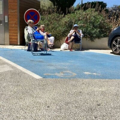 Drei ältere Damen sizen in Campingstühlen auf einem Parkplatz, die Sonne scheint.